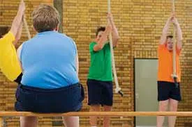 Obese boy sitting on the bench watching other boys climb ropes in gym class